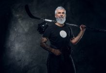 Tattooed old man athlete with hockey stick against dark background