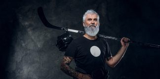 Tattooed old man athlete with hockey stick against dark background